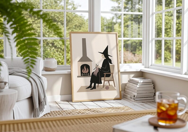 Op de afbeelding: Een ingelijste kunstdruk met een heks die bij een open haard breit. De illustratie is in minimalistische stijl met een neutraal kleurenpalet. De heks draagt een zwarte jurk en hoed. Het kunstwerk wordt getoond in een lichte kamer.