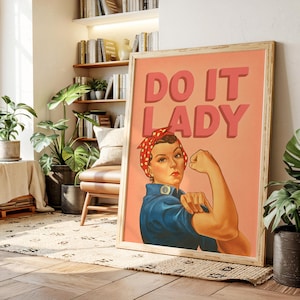 Può includere: Un poster incorniciato con la scritta "DO IT LADY" in lettere rosse. L'immagine presenta una donna con una camicia blu e un foulard a pois rossi che flette il braccio. Il poster è in una cornice di legno chiaro.
