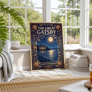 May include: A framed print of "The Great Gatsby" by F. Scott Fitzgerald. The artwork features a night scene with a mansion, moon, and water, framed by gold floral accents. The print is leaning against a window, with a stack of books nearby.