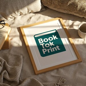 May include: A framed print with the text "Book Tok Print" in white on a teal green book icon. The frame is light brown. Beside the frame are an open book, glasses, and a ceramic mug.