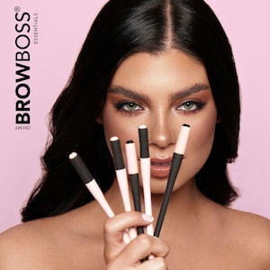 May include: A woman holds five black and pink brow pencils with a white tip. The pencils are held in front of her face. The background is pink. The text "BROWBOSS® ESSENTIALS" is in the upper left corner of the image.