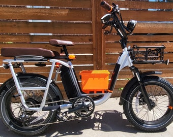Puede incluir: Una bicicleta eléctrica plateada y negra con un asiento marrón, una cesta negra y una batería naranja. La bicicleta está aparcada delante de una valla de madera.