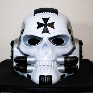 May include: A white skull helmet with a black cross on the forehead. The helmet has black and silver details and a black and silver breathing apparatus.
