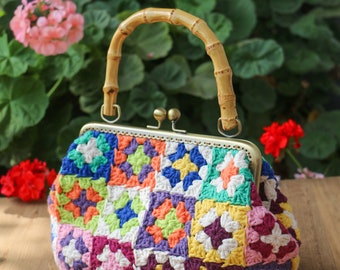 May include: A colorful crocheted handbag with a bamboo handle and a brass clasp. The bag is made of granny squares in various colors, including pink, purple, green, yellow, and orange.