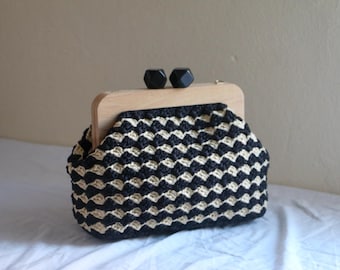 Könnte beinhalten: Handgefertigte Clutch mit einem schwarz-cremefarbenen Häkelmuster. Die Tasche hat einen Holzrahmen mit zwei schwarzen geometrischen Verschlüssen. Die Clutch liegt auf einer weißen Oberfläche.
