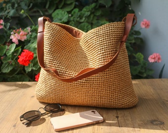 May include: A woven straw tote bag with brown leather straps. The bag is tan and has a spacious interior. Sunglasses and a smartphone are on a wooden surface in front of the bag. Flowers and greenery are in the background.