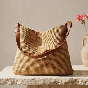 May include: A tan woven hobo bag featuring a textured, grid-like pattern and a smooth brown leather shoulder strap. The bag has a wide, open top design with a structured base, showcasing a consistent, uniform weave throughout the exterior surface.