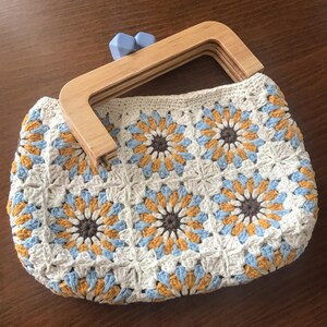 May include: A handmade crochet handbag with a wooden handle. The bag features a floral pattern in white, yellow, light blue, and brown. The wooden handle is light-colored, and the clasp is a light blue color.