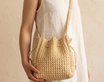 May include: A beige crochet bucket bag with a textured, woven pattern and a drawstring closure. The bag features a long, matching shoulder strap and is constructed from a light-colored, fibrous material with a consistent, repeating stitch design.