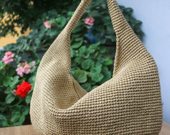 May include: A beige hobo-style shoulder bag crafted from a dense, textured crochet knit. The bag features a single integrated handle that curves upward from the main body, creating a slouchy, crescent-shaped silhouette with a wide, open top.