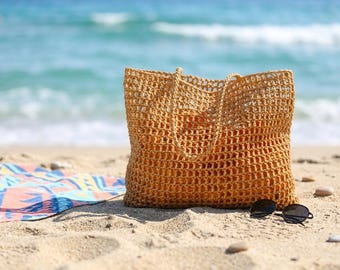 May include: A rectangular, golden-brown crochet tote bag with an open-weave grid pattern and two matching shoulder straps. The bag is positioned on a sandy surface next to a pair of black-framed sunglasses and a colorful patterned fabric.