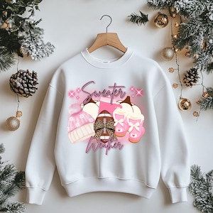 Puede incluir: Sudadera blanca con un diseño gráfico que presenta un gorro de punto rosa, un balón de fútbol americano, calabazas y botas. Las palabras "Sweater Weather" están escritas en rosa. El diseño tiene un tema de otoño o invierno.