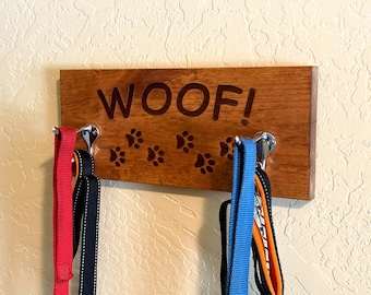 May include: Wooden dog leash rack with the word "WOOF!" engraved on it. The rack has two hooks and is decorated with paw prints. Three leashes are hanging on the hooks, one red, one black, and one blue.
