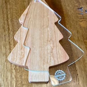 May include: A wooden cutting board in the shape of a Christmas tree, with a clear acrylic overlay. The wood has a light brown color and a visible grain. The acrylic overlay has a logo in the bottom right corner.