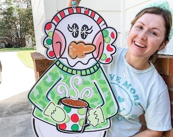May include: A snowman holding a green and red mug with a brown liquid inside. The snowman is wearing a green and brown sweater with a leopard print. The snowman has a red and white hat and a carrot nose. The snowman is standing on a green and white striped base. The snowman is holding a red and white mug with a brown liquid inside. The snowman is wearing a green and brown sweater with a leopard print. The snowman has a red and white hat and a carrot nose. The snowman is standing on a green and white striped base.
