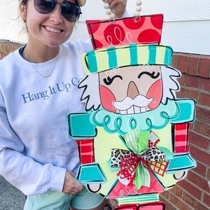 May include: A colorful wooden nutcracker door hanger with a red hat, green and blue body, and a red bow. The nutcracker is holding a wooden bead garland.