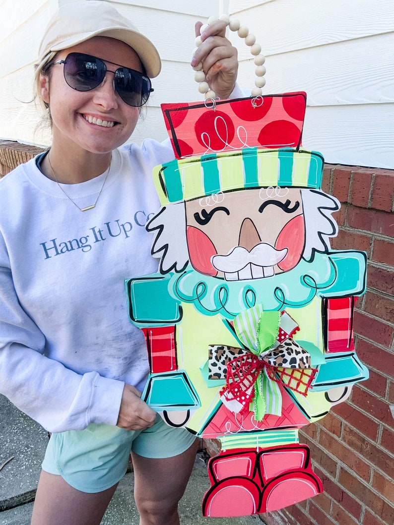 May include: A colorful wooden nutcracker door hanger with a red hat, green and blue body, and a red bow. The nutcracker is holding a wooden bead garland.