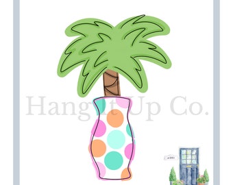 May include: A green palm tree in a pink and white polka dot vase with orange and turquoise dots. The vase is sitting on a white background with the text "Hang It Up Co." in black.