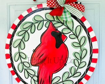 May include: A round, red and white striped door hanger with a black and white border. A red cardinal bird is painted in the center of the hanger, surrounded by green leaves. A red, white, and green bow is attached to the top of the hanger.