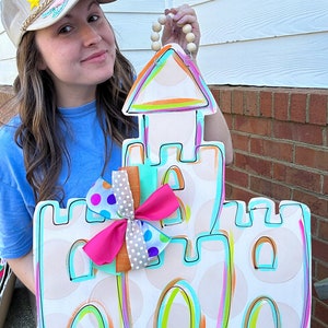 May include: A white wooden sandcastle door hanger with a pink, blue, and white polka dot bow. The sandcastle is painted with bright colors, including turquoise, pink, orange, and yellow. The door hanger has a wooden bead loop for hanging.