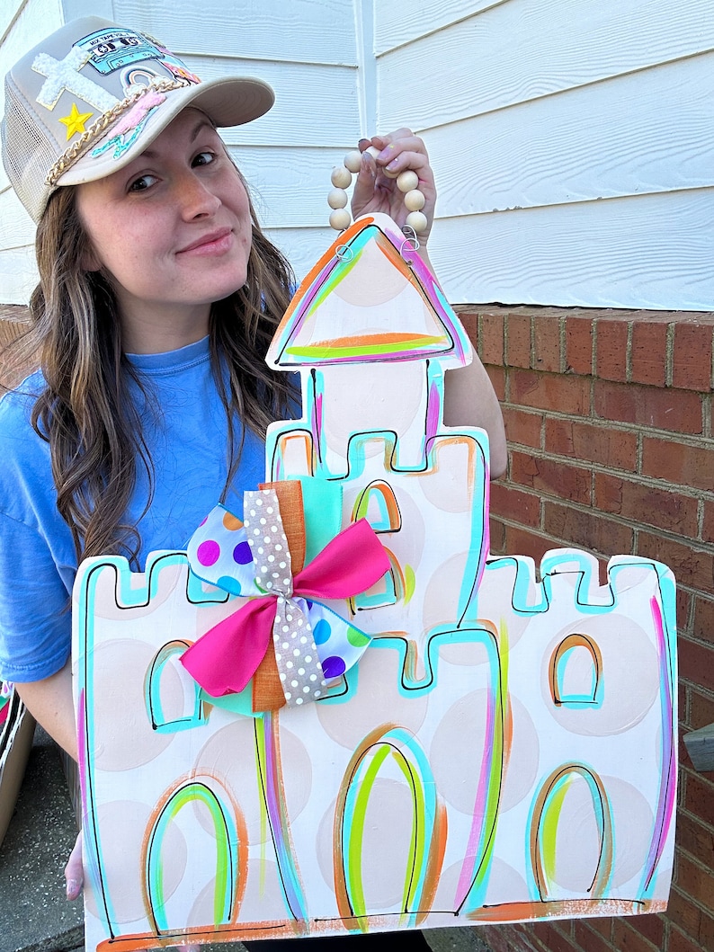 May include: A white wooden sandcastle door hanger with a pink, blue, and white polka dot bow. The sandcastle is painted with bright colors, including turquoise, pink, orange, and yellow. The door hanger has a wooden bead loop for hanging.
