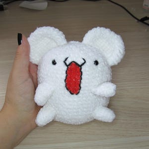 Puede incluir: Un peluche blanco con forma de personaje de dibujos animados, con orejas grandes y redondas y una lengua roja. El juguete tiene pequeños ojos negros y se sostiene en una mano.