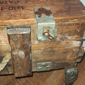 May include: A wooden box with a metal latch and a rope handle. The box is sitting on a wooden floor and has the text "1910-1" and "3 PATR" printed on the side.