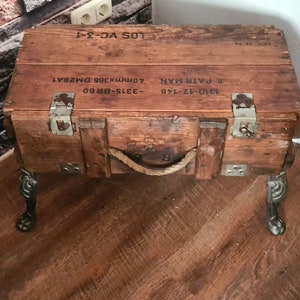 May include: A wooden box with metal hardware and a rope handle, repurposed as a side table with black metal legs. The box has printed text on the top: "310-12-140 E. FAIR-HAWK  ADM173019E  DMB541  LOS VC-3-1"