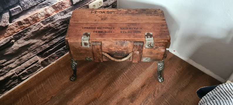 May include: A wooden box with metal hardware and a rope handle, repurposed as a side table with black metal legs. The box has printed text on the top: "310-12-140 E. FAIR-HAWK  ADM173019E  DMB541  LOS VC-3-1"