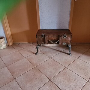 May include: A wooden storage trunk with metal accents and rope detailing, used as a table with four metal legs. The trunk is brown and has a distressed finish.