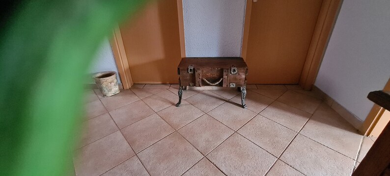 May include: A wooden storage trunk with metal accents and rope detailing, used as a table with four metal legs. The trunk is brown and has a distressed finish.