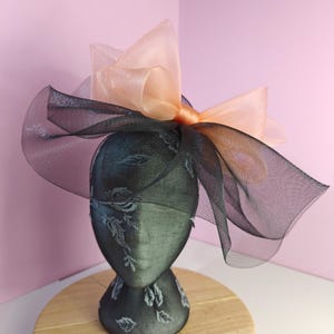 Könnte beinhalten: Dekoratives Kopfstück mit schwarzem Mesh-Rand und großer pfirsichfarbener Schleife. Das Kopfstück wird auf einem Mannequinkopf mit floralem Muster präsentiert. Der Hintergrund ist ein zartes Rosa.