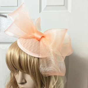 Könnte beinhalten: Ein peachfarbener Fascinator mit einer großen Schleife aus Netz. Der Fascinator wird auf dem Kopf einer Mannequin getragen.