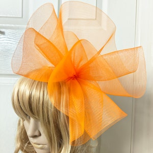 Könnte beinhalten: Ein orangefarbener Fascinator aus Netzstoff, ein Kopfschmuck mit großem Schleifendesign. Der Kopfschmuck besteht aus einem transparenten, gewebten Material und ist an einer Haarspange befestigt. Der Kopfschmuck ist leuchtend orange und wird auf einem Schaufensterkopf mit blonden Haaren getragen.