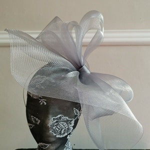 May include: A silver mesh fascinator with a large bow. The fascinator is on a black and white floral mannequin head.