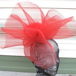 May include: A bright red fascinator, crafted from layers of delicate, transparent material. The headpiece is adorned with a central cluster of feathers. The base is a black head form with a floral design, offering a bold contrast.