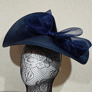 May include: A navy blue wide-brimmed hat with a large bow. The hat is made of woven material and has a structured shape. The bow is made of a sheer fabric. The hat is displayed on a mannequin head.