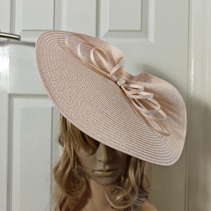 Könnte beinhalten: Ein breitkrempiger Hut aus Stroh in hellrosa. Der Hut hat eine große Schleife an der Seite und wird von einer Puppe mit langen blonden Haaren getragen.