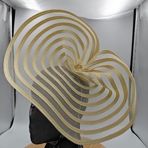 May include: A striking beige hat with a distinctive, layered design. The hat has a wide brim and features a series of horizontal, curved bands, creating an openwork effect. It is displayed on a black mannequin head, highlighting its shape and structure.