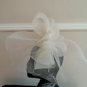 May include: A white tulle fascinator with a large bow. The fascinator is on a black mannequin head with a floral pattern.