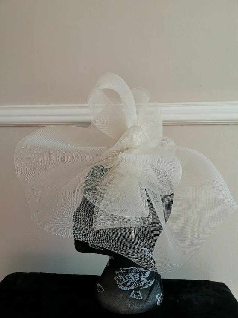 May include: A white tulle fascinator with a large bow. The fascinator is on a black mannequin head with a floral pattern.