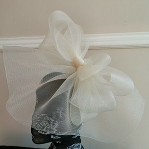 May include: A large white tulle bow with a gathered center. The bow is on a mannequin head.
