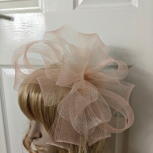 Könnte beinhalten: Hellrosa Kopfschmuck mit einem großen, mehrschichtigen Schleifendesign. Das Accessoire besteht aus einem transparenten, strukturierten Stoff. Präsentiert auf einem Schaufensterkopf mit blonden Haaren. Geeignet für besondere Anlässe.