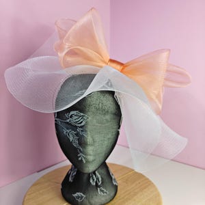 Könnte beinhalten: Dekoratives Kopfstück mit einer schwarzen Kopfform mit floralem Design. Das Kopfstück hat eine große pfirsichfarbene Schleife und einen weißen Netzschleier. Die Kopfform steht auf einem Holzständer vor einem rosa Hintergrund.