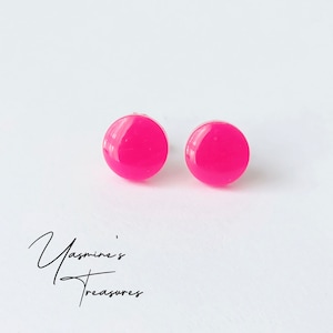 May include: A pair of bright pink, round stud earrings. The earrings are glossy and appear to be made of a solid material. The earrings are set against a white background. The text "Yasmine's Treasures" is in the lower left corner.