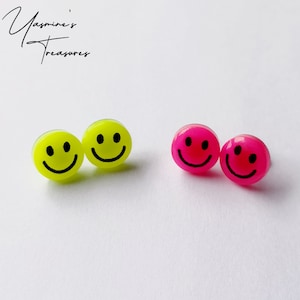Peut inclure: Deux paires de boucles d'oreilles avec des smileys. Une paire est jaune avec des smileys noirs et l'autre paire est rose avec des smileys noirs.