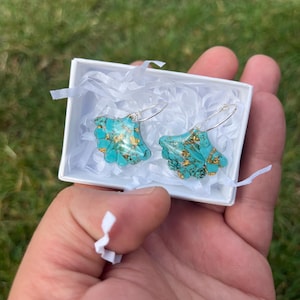 Op de afbeelding: Een paar turquoise oorbellen met gouden accenten, gepresenteerd in een witte geschenkdoos met opvulmateriaal. De oorbellen hebben een unieke, organische vorm en zijn bevestigd aan zilverkleurige oorhaken. De doos wordt in een hand gehouden.