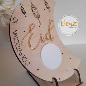 Peut inclure: Compteur de jours jusqu'à l'Aïd en forme de croissant de lune en bois avec inscription dorée et un cercle blanc au centre. Le panneau est doté d'un support et est décoré d'étoiles et de lanternes.