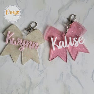 May include: Two bow-shaped keychains with silver-toned metal lobster clasps. One keychain is translucent beige with the name Kwynn in pink script. The other is translucent pink with glitter and the name Kalise in white script lettering.