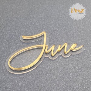 Puede incluir: Corte de acrílico dorado de la palabra "junio" con fondo transparente. El corte está en capas, creando un efecto 3D.